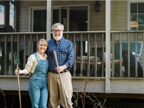 A couple standing outside their home holding garden tools